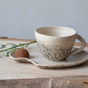 May include: A white ceramic teacup and saucer with a speckled gray glaze. A single chocolate truffle sits on the saucer.