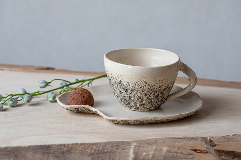 May include: A white ceramic teacup and saucer with a speckled gray glaze. A single chocolate truffle sits on the saucer.