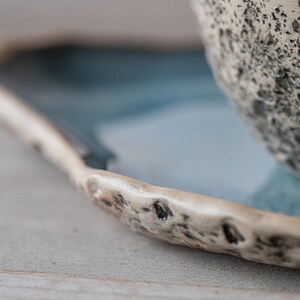 May include: A close-up of a ceramic saucer with a blue glaze and a textured rim. The saucer is partially filled with a white ceramic bowl with a textured surface.