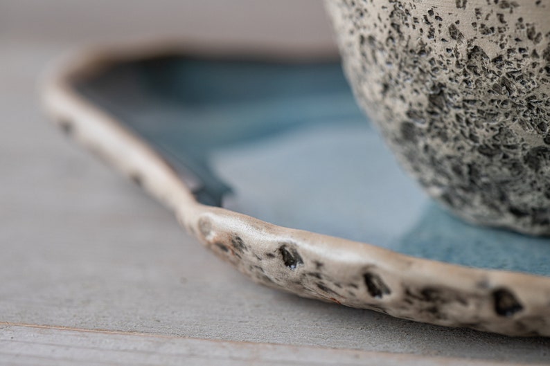 May include: A close-up of a ceramic saucer with a blue glaze and a textured rim. The saucer is partially filled with a white ceramic bowl with a textured surface.