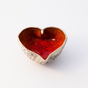 May include: A heart-shaped ceramic bowl with a white exterior and a red and orange interior glaze.