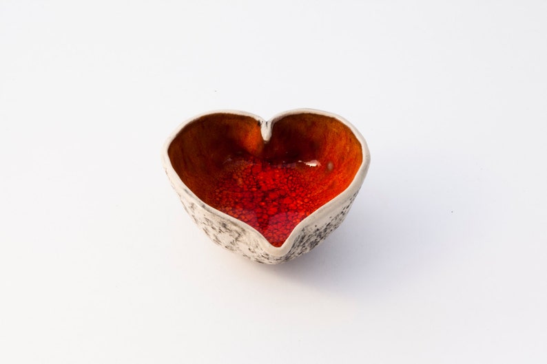 May include: A heart-shaped ceramic bowl with a white exterior and a red and orange interior glaze.