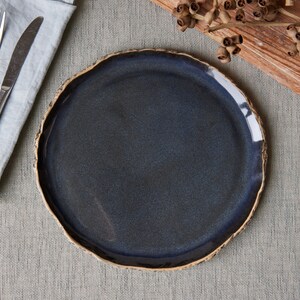 May include: A round ceramic dinner plate with a dark blue, glossy glazed center and a textured, uneven light brown rim. The plate features a deep indigo hue that transitions into a lighter blue tone, showcasing a handcrafted, organic edge design.