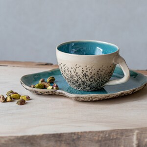 May include: A teal and white ceramic teacup with a speckled design sits on a teal saucer. The saucer has an irregular shape and is on a wooden surface. A small pile of pistachios is in the foreground.