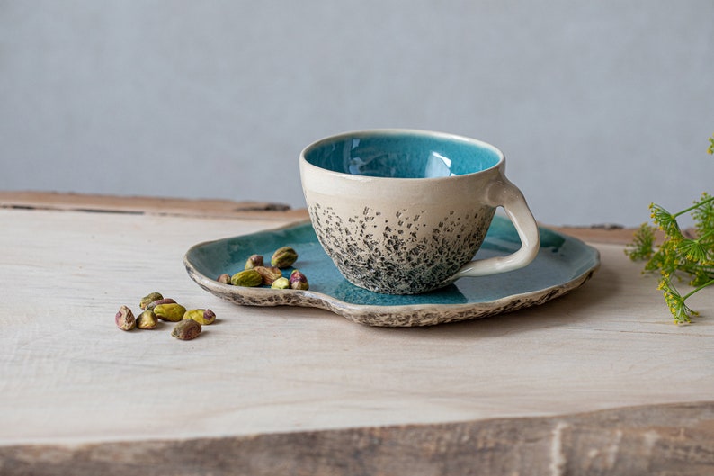 May include: A teal and white ceramic teacup with a speckled design sits on a teal saucer. The saucer has an irregular shape and is on a wooden surface. A small pile of pistachios is in the foreground.