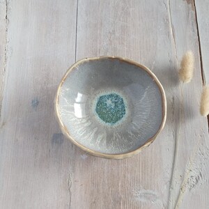May include: A handmade ceramic bowl with a unique design. The bowl features a light grey exterior with a teal and green centre. The rim has a gold-coloured trim. The bowl is approximately 15 cm in diameter.