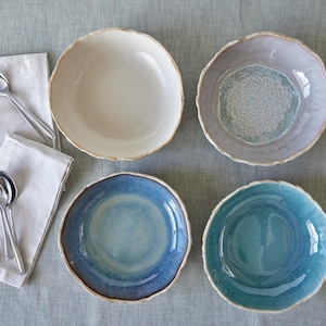 May include: Four ceramic bowls in different colors and patterns. The bowls are arranged in a circle on a light gray surface. The bowls are all round and have a slightly irregular rim.