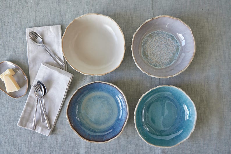 May include: Four ceramic bowls in different colors and patterns. The bowls are arranged in a circle on a light gray surface. The bowls are all round and have a slightly irregular rim.
