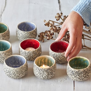 Könnte beinhalten: Ein Set aus sieben kleinen, weißen Kerzenhaltern aus Keramik mit strukturierten Oberflächen. Die Kerzenhalter sind in einem Kreis auf einer weißen Holzoberfläche angeordnet. Einige der Kerzenhalter haben türkisfarbene, blaue oder rote Innenräume, und zwei der Kerzenhalter haben brennende Teelichter im Inneren.
