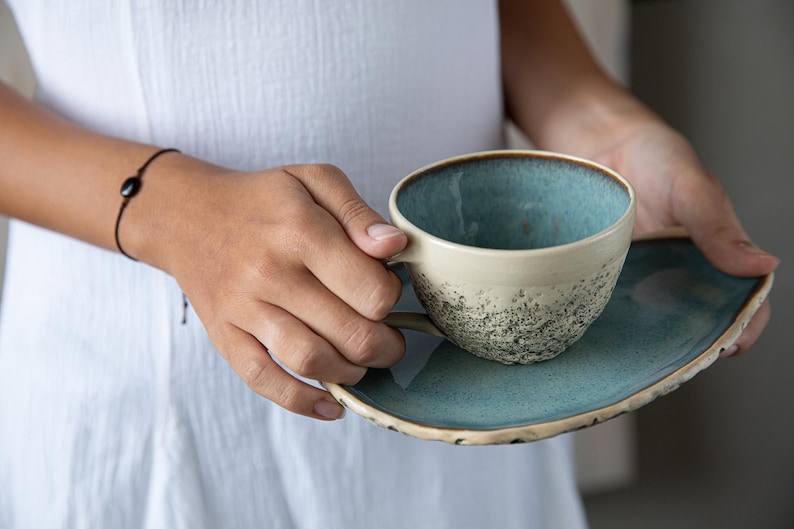 May include: A blue and white ceramic teacup and saucer with a speckled glaze. The cup has a rounded shape and a handle. The saucer is a simple, rounded shape.