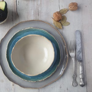 May include: A ceramic dinnerware set consisting of a large grey plate, a medium blue plate, and a small cream-colored bowl. A silver fork and a silver knife with ornate handles are placed to the right. An avocado and walnuts are nearby.