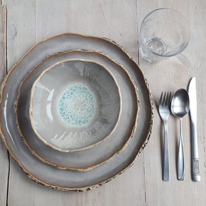 May include: A ceramic dinnerware set consisting of a large plate, a smaller plate, and a bowl, all featuring a grey glaze with a textured, uneven brown rim. A clear glass, a silver fork, a silver spoon, and a silver knife are arranged beside the plates.