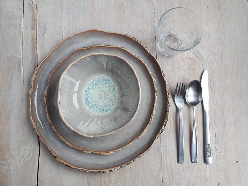 May include: A ceramic dinnerware set consisting of a large plate, a smaller plate, and a bowl, all featuring a grey glaze with a textured, uneven brown rim. A clear glass, a silver fork, a silver spoon, and a silver knife are arranged beside the plates.