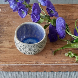 May include: A small, blue and white ceramic bowl with a textured exterior sits on a wooden cutting board. A bouquet of purple flowers with green stems is placed next to the bowl.