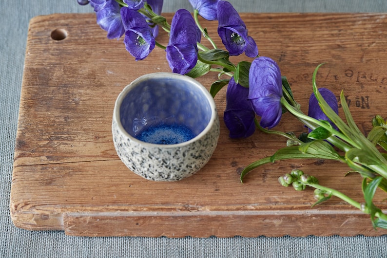 May include: A small, blue and white ceramic bowl with a textured exterior sits on a wooden cutting board. A bouquet of purple flowers with green stems is placed next to the bowl.