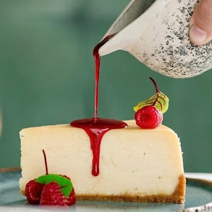 Puede incluir: Una porci&oacute;n de tarta de queso cubierta con una salsa roja y dos frambuesas. La salsa se vierte desde una jarra blanca.