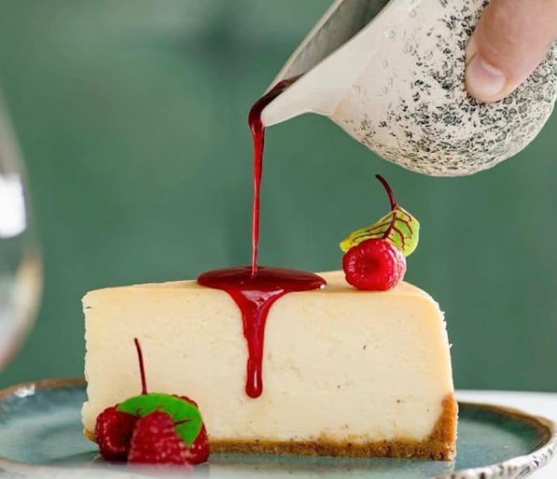 Puede incluir: Una porci&oacute;n de tarta de queso cubierta con una salsa roja y dos frambuesas. La salsa se vierte desde una jarra blanca.