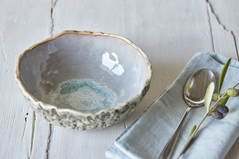 May include: A gray ceramic bowl with a textured exterior and a blue and white glaze inside. The bowl is sitting on a white wooden surface.
