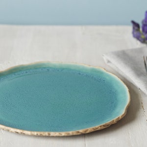 May include: A round ceramic dinner plate with a vibrant teal blue glaze and a textured, uneven beige rim. The surface of the plate features a subtle speckled finish, while the edges have a natural, organic shape that contrasts with the smooth center.