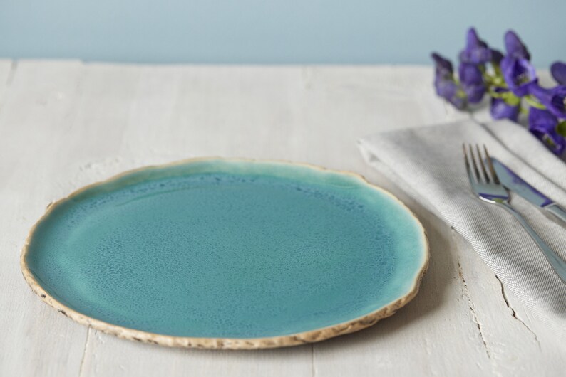 May include: A round ceramic dinner plate with a vibrant teal blue glaze and a textured, uneven beige rim. The surface of the plate features a subtle speckled finish, while the edges have a natural, organic shape that contrasts with the smooth center.