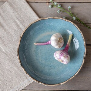 May include: Two whole garlic bulbs with purple stems on a blue ceramic serving bowl. The bowl has a white rim and is on a light brown wooden surface.
