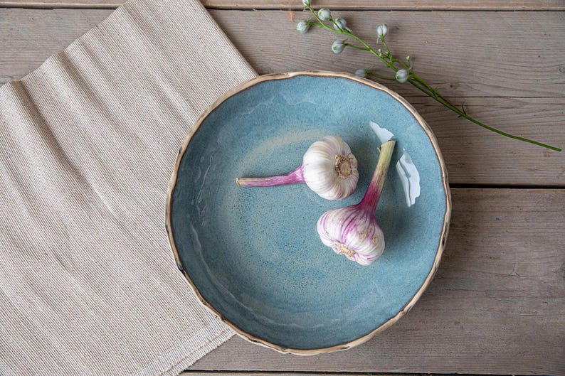 May include: Two whole garlic bulbs with purple stems on a blue ceramic serving bowl. The bowl has a white rim and is on a light brown wooden surface.