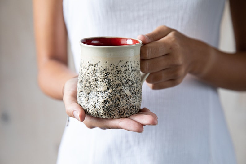 May include: A ceramic mug with a textured, speckled surface in shades of gray and white. The mug has a red rim and a handle.