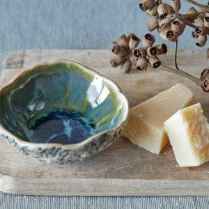 May include: A small, blue and green ceramic bowl with a textured rim sits on a wooden cutting board with two blocks of white soap.