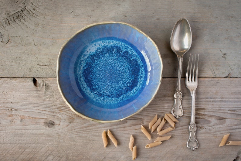 May include: A blue ceramic bowl with a white and blue speckled design sits on a wooden surface with a silver spoon and fork beside it.  A few pieces of pasta are scattered around the bowl.