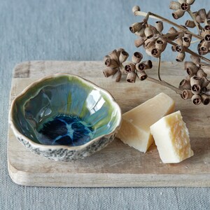 K&ouml;nnte beinhalten: Eine kleine, blaue und gr&uuml;ne Keramiksch&uuml;ssel mit strukturiertem Rand steht auf einem Holzbrett mit zwei St&uuml;cken wei&szlig;em K&auml;se und einem Zweig getrocknetem Eukalyptus.