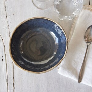 May include: A dark blue ceramic bowl with a slightly rippled rim. The bowl is empty and sits on a white wooden surface.