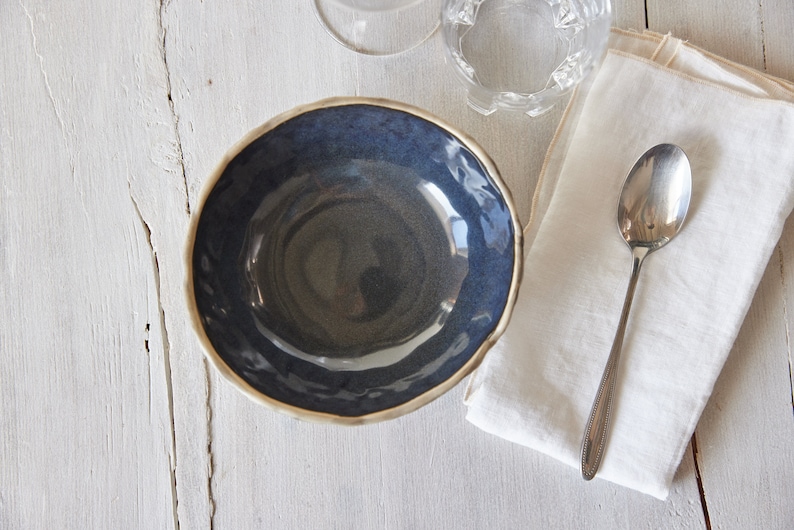 May include: A dark blue ceramic bowl with a slightly rippled rim. The bowl is empty and sits on a white wooden surface.