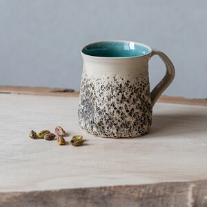 May include: A speckled ceramic mug with a light beige body, a dark gray speckled pattern, and a teal blue interior. The mug is sitting on a light wood surface.
