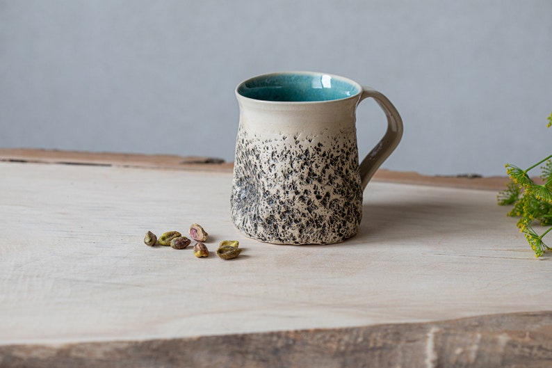 May include: A speckled ceramic mug with a light beige body, a dark gray speckled pattern, and a teal blue interior. The mug is sitting on a light wood surface.