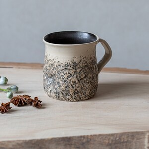 May include: A ceramic mug with a textured, speckled black and white glaze. The mug has a rounded body and a handle. The mug is sitting on a wooden surface with star anise pods.