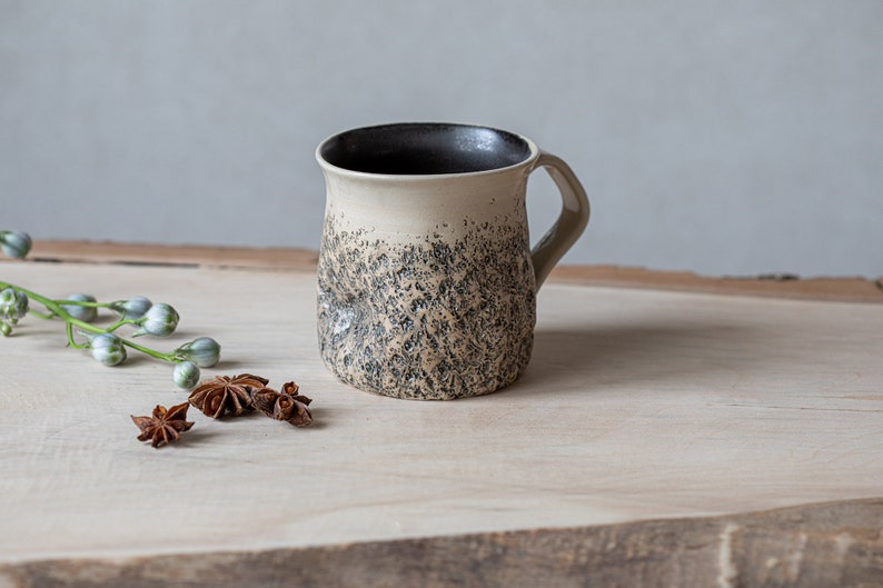 May include: A ceramic mug with a textured, speckled black and white glaze. The mug has a rounded body and a handle. The mug is sitting on a wooden surface with star anise pods.