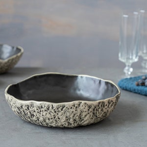 May include: A set of three ceramic bowls with a speckled, textured finish. The bowls are black on the inside and have a light gray exterior with black speckles. The bowls are arranged on a gray surface.