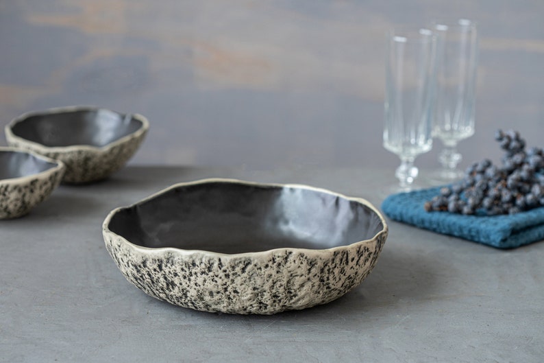 May include: A set of three ceramic bowls with a speckled, textured finish. The bowls are black on the inside and have a light gray exterior with black speckles. The bowls are arranged on a gray surface.