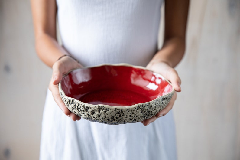 May include: A red ceramic bowl with a textured, speckled rim. The bowl is held by a person's hands.
