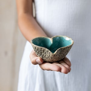 Puede incluir: Un cuenco de cer&aacute;mica en forma de coraz&oacute;n con un exterior texturizado y un glaseado azul verdoso en el interior. El cuenco est&aacute; siendo sostenido en la mano de una persona.