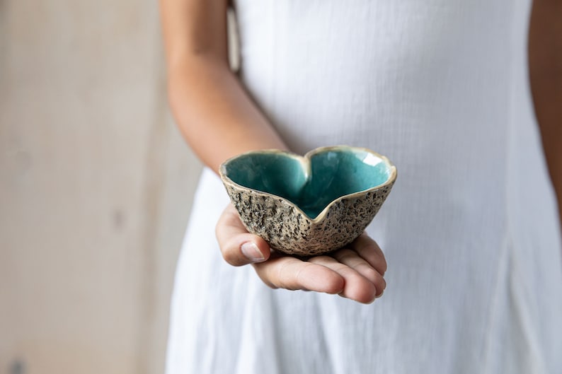 Puede incluir: Un cuenco de cer&aacute;mica en forma de coraz&oacute;n con un exterior texturizado y un glaseado azul verdoso en el interior. El cuenco est&aacute; siendo sostenido en la mano de una persona.