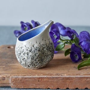 Puede incluir: Un peque&ntilde;o creamer de cer&aacute;mica blanca con un glaseado azul en el interior y un exterior texturizado moteado. El creamer est&aacute; sentado sobre una tabla de cortar de madera junto a una rama de flores moradas.