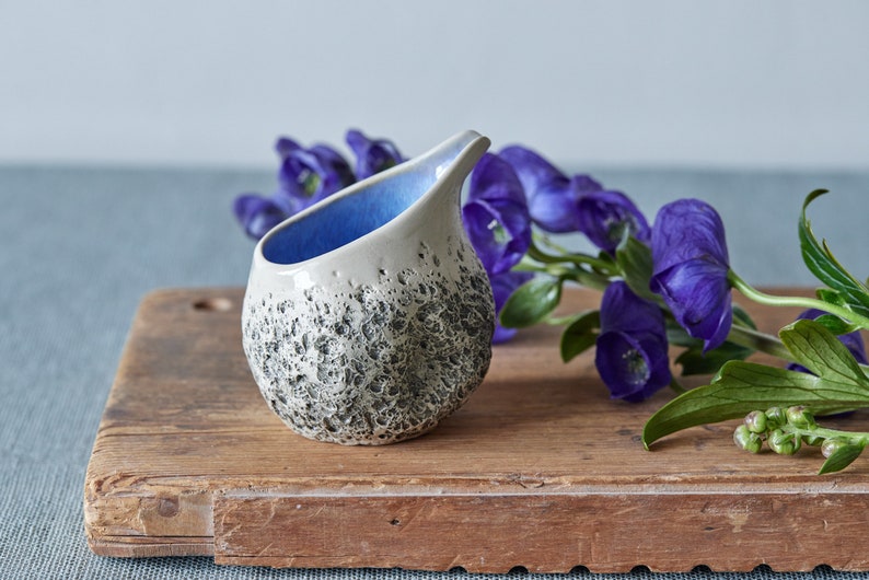 Puede incluir: Un peque&ntilde;o creamer de cer&aacute;mica blanca con un glaseado azul en el interior y un exterior texturizado moteado. El creamer est&aacute; sentado sobre una tabla de cortar de madera junto a una rama de flores moradas.