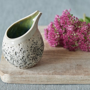 Puede incluir: Un peque&ntilde;o creamer de cer&aacute;mica blanca con una superficie texturizada moteada y un glaseado verde en el interior. El creamer tiene forma de l&aacute;grima y tiene un peque&ntilde;o pico. Est&aacute; sentado en una tabla de cortar de madera junto a un ramo de flores rosas.