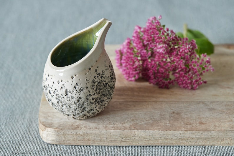 Puede incluir: Un peque&ntilde;o creamer de cer&aacute;mica blanca con una superficie texturizada moteada y un glaseado verde en el interior. El creamer tiene forma de l&aacute;grima y tiene un peque&ntilde;o pico. Est&aacute; sentado en una tabla de cortar de madera junto a un ramo de flores rosas.