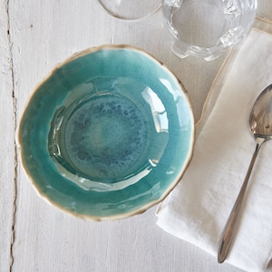 May include: A teal blue ceramic bowl with a slightly rippled edge sits on a white wooden surface. A white linen napkin and a silver spoon are to the right of the bowl.