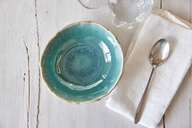 May include: A teal blue ceramic bowl with a slightly rippled edge sits on a white wooden surface. A white linen napkin and a silver spoon are to the right of the bowl.