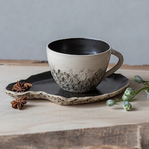 May include: A white ceramic coffee cup with a black interior and black speckles on the exterior, sitting on a black ceramic saucer with a wavy edge. The cup and saucer are on a light brown wooden surface with star anise pods and a green stem with small blue flowers.