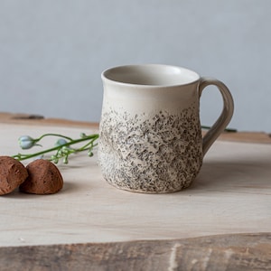 May include: A white ceramic mug with a speckled brown glaze. The mug has a rounded body and a handle. Two chocolate truffles are in the foreground.