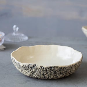 May include: A white ceramic bowl with a speckled black and gray glaze. The bowl has a wide, shallow shape and a slightly rippled edge.